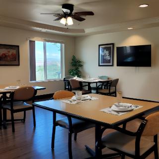 A dining area with tables and chairs, featuring a ceiling fan and a television, illuminated by natural light from the window.