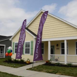 A suburban yellow house with promotional flags and balloons outside, likely for a real estate event.