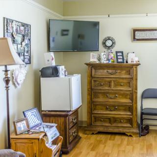 A cozy living room corner featuring a wall-mounted television, a wooden dresser, and various decorations including framed pictures and a lamp.