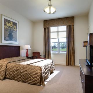 A well-lit hotel bedroom featuring a large bed, bedside lamp, and a window with curtains.