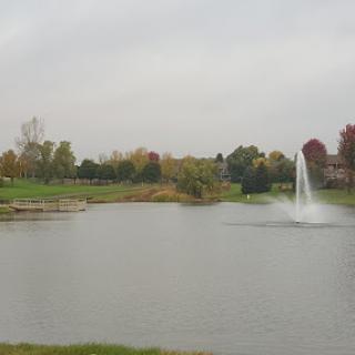 A serene view of a lake with a fountain, surrounded by autumn foliage on a cloudy day.