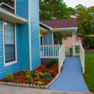 A house with blue siding features a wheelchair-accessible ramp leading to the porch, surrounded by a landscaped garden with colorful flowers.