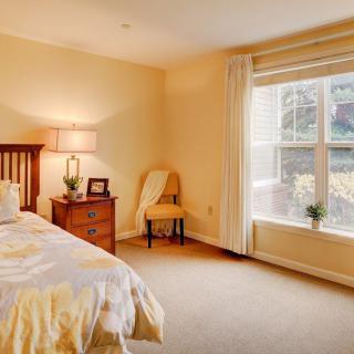 A cozy bedroom featuring a bed, wooden nightstand with a lamp, and a large window letting in natural light.