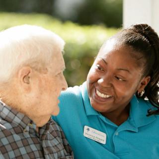 A caregiver smiling and providing support to an elderly person outdoors.