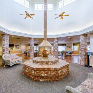 A spacious lobby featuring a central brick fireplace surrounded by seating, with ceiling fans and ample natural lighting.