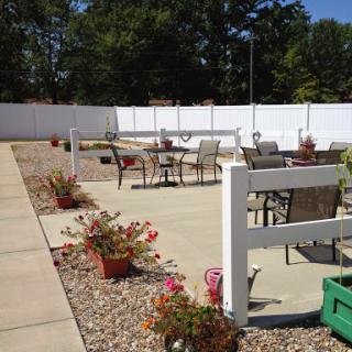 A sunny outdoor patio area with seating and blooming flowers, surrounded by a white fence.
