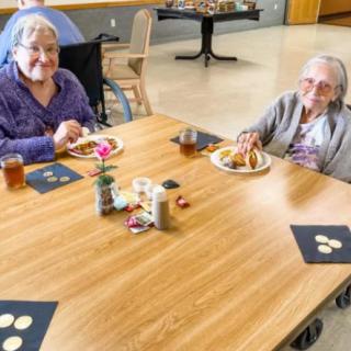 Two elderly women enjoying a meal together at a dining table indoors.