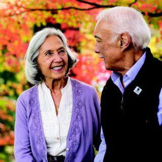 An elderly couple smiling at each other against a backdrop of autumn leaves.