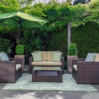 A cozy outdoor patio setup with wicker furniture, cushions, a table, and an umbrella surrounded by lush greenery.