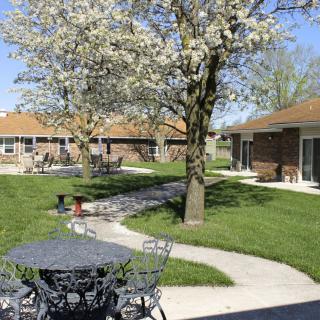 A peaceful courtyard with blossoming trees, outdoor seating, and a winding pathway amidst well-kept lawns and surrounding buildings.