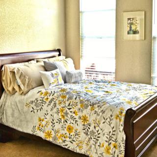 A cozy bedroom featuring a wooden bed with floral bedding and multiple pillows, lit by natural sunlight from a nearby window.