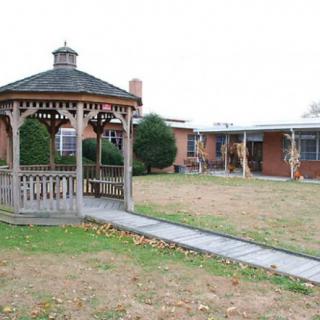 A wooden gazebo stands in a yard next to a brick building, with a paved pathway leading to it.
