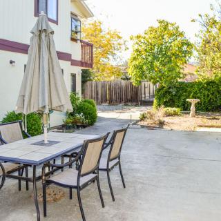 A sunny backyard with a patio table, chairs, and an umbrella surrounded by greenery.