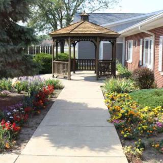 A sunny pathway leads to a wooden gazebo surrounded by vibrant flowers and well-maintained landscaping.