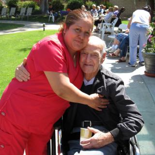 A caregiver hugging an elderly person in a wheelchair outdoors.