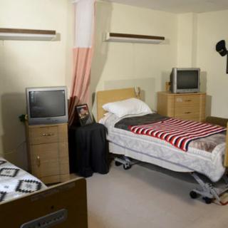 A hospital room with two beds, each equipped with a television and bedside furniture.
