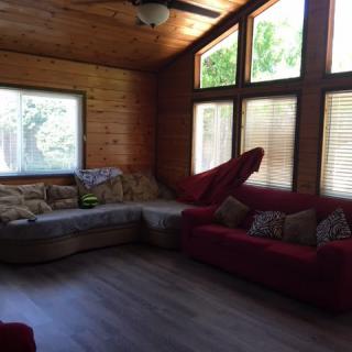 A cozy living room with wood paneling, featuring a sectional sofa and a red couch near large windows.