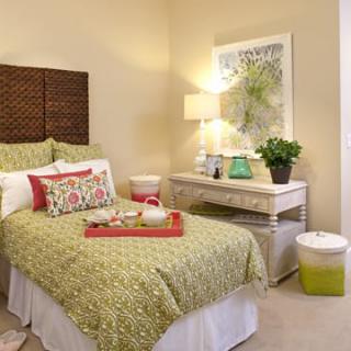 A cozy bedroom with warm decor, featuring a bed with green bedding, a small desk with a lamp, and a decorative plant.
