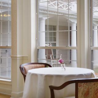 An elegant dining area with a round table, set for two, by large windows letting in natural light.