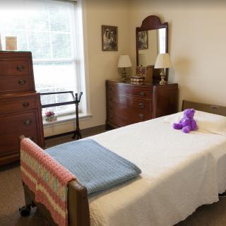 A cozy bedroom featuring a bed with a purple teddy bear, dressers with mirrors, and a window letting in natural light.