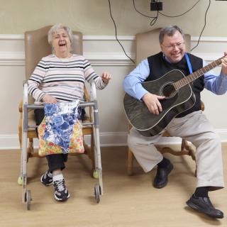 An elderly woman in a wheelchair enjoys listening to a man playing the guitar, both smiling and sharing a joyful moment.