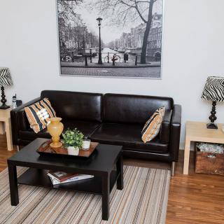 A cozy living room with a dark leather sofa, accented by pillows and flanked by stylish lamps on end tables. A black coffee table holds decorative items and a plant, while a large framed artwork hangs above the sofa. A striped rug adds texture to the wood