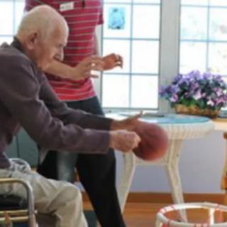 An elderly person in a wheelchair playing basketball indoors, assisted by a caregiver.