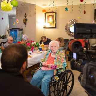 An elderly woman in a wheelchair being interviewed during a birthday celebration, surrounded by festive decorations.