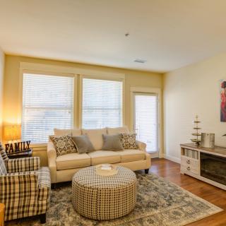 A cozy living room with a sofa, armchair, and ottoman arranged around a patterned carpet. Natural light filters through the large windows, creating a warm and inviting atmosphere.