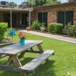 A picnic table is set on a lawn in front of a brick building, surrounded by lush greenery and flowers.