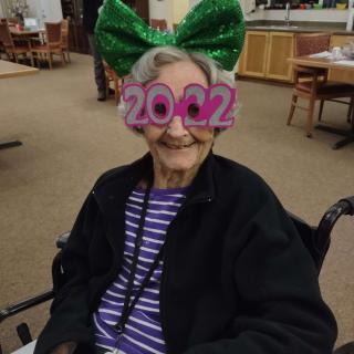 An elderly person in a wheelchair wearing festive 2022 glasses and a large green bow.