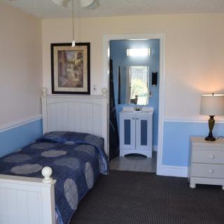 A cozy bedroom with a single bed, featuring blue and white decor, a nightstand with a lamp, and an adjoining bathroom.