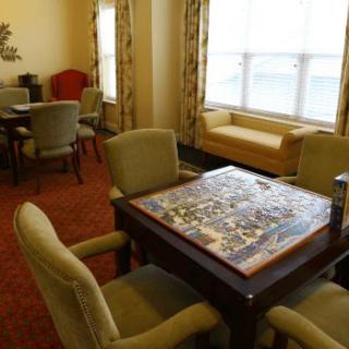 A cozy living room with a puzzle laid out on a table surrounded by chairs, next to a large window.