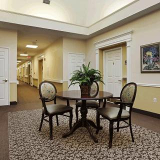 A warmly lit hallway with a table and chairs, decorated with a potted plant, surrounded by carpeted flooring and artwork on the wall.