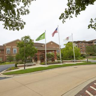 A building with three flags in front, surrounded by trees and a driveway on a cloudy day.