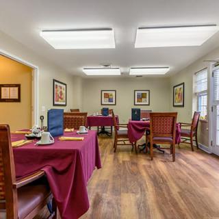 A cozy dining room with tables set for service, featuring wooden floors and artwork on the walls.