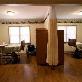 A shared nursing home room with two hospital beds separated by a curtain, featuring a wheelchair and wooden flooring.