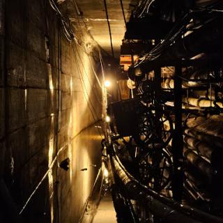 An underground tunnel illuminated by artificial light, showcasing industrial elements and water reflections.