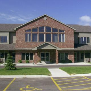 A modern brick building with a large entrance, flanked by numerous windows, and a front parking lot under a clear blue sky.