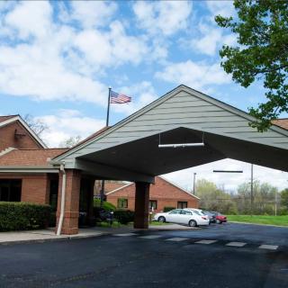 A brick building with a covered entrance and driveway, featuring an American flag and surrounded by trees under a cloudy sky.