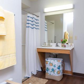 A well-lit bathroom with a sink, mirror, and shower area adorned with yellow towels and a colorful laundry basket.