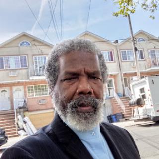 A man with a beard poses for a selfie in front of residential buildings on a sunny day.