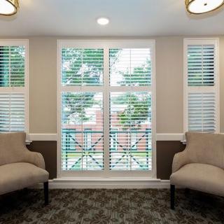 A cozy seating area with two chairs facing large windows with blinds, carpeted flooring, and ceiling lights.