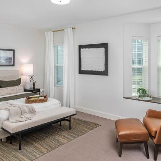 A modern minimalist bedroom with neutral tones, featuring a bed, chair, and window.