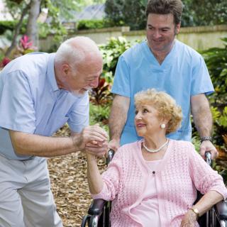 An elderly woman in a wheelchair is being attended to by a nurse and interacting with an older man in an outdoor setting.