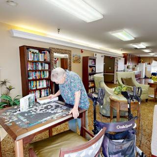 A senior individual engaged in a puzzle activity in a cozy, well-lit community space with bookshelves and comfortable seating.