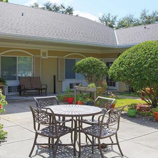 A sunny patio area featuring a round table with chairs surrounded by greenery and well-maintained bushes.