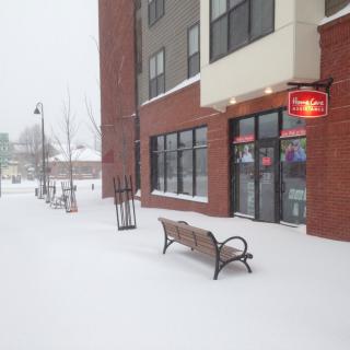 A snowy street scene with a bench and a building.