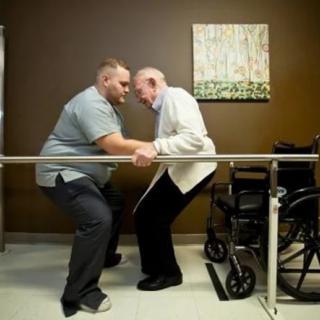 A caregiver assists an elderly man with physical therapy exercises using parallel bars.