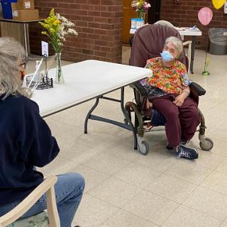 An elderly woman in a wheelchair engages in conversation with another person, both wearing masks during a social visit.
